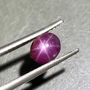 Könnte beinhalten: Ein tief magentafarbener Sternrubin-Edelstein mit einem glatten, polierten Cabochon-Schliff. Der Stein zeigt einen ausgeprägten sechsstrahligen Sterneffekt, bekannt als Asterismus, der auf seiner Oberfläche zentriert ist. Der Stein wird von einer silberfarbenen Pinzette gehalten.