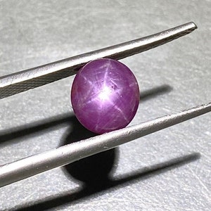 Könnte beinhalten: Ein runder, im Cabochon-Schliff gefertigter Sternrubin-Edelstein mit einem tiefen Magenta-Violett-Ton. Der Stein zeigt einen ausgeprägten sechsstrahligen Asterismus-Effekt, der durch ein helles, sich kreuzendes Sternmuster in der Mitte entsteht.