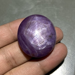 May include: A polished, oval-shaped gemstone with a deep purple hue. The stone has a star-like effect, with lines radiating from the center. The surface reflects light, creating a shimmering appearance. The gemstone is held in a hand.