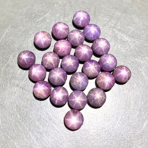 Könnte beinhalten: Eine Sammlung runder, polierter Edelsteine in verschiedenen Lilatönen. Jeder Stein zeigt ein sternförmiges Muster auf seiner Oberfläche. Die Edelsteine sind auf einer grauen Oberfläche verstreut.