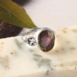 May include: A silver ring with a large, oval-cut, deep red gemstone. The ring has decorative detailing on the band and around the stone setting. The gemstone has a slightly translucent appearance. The ring is set on a white and gray surface.