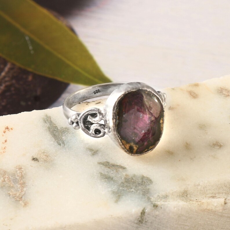 May include: A silver ring with a large, oval-cut, deep red gemstone. The ring has decorative detailing on the band and around the stone setting. The gemstone has a slightly translucent appearance. The ring is set on a white and gray surface.