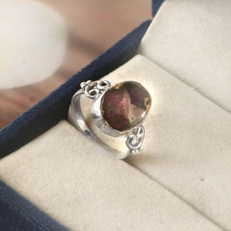 May include: A silver ring featuring an oval, dark red gemstone. The ring has decorative swirls on the sides and is set in a silver band. The ring is presented in a blue jewellery box.