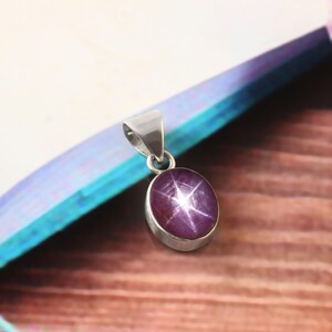 May include: A silver pendant featuring an oval, purplish-red gemstone with a six-rayed star pattern. The gemstone is set in a silver bezel and has a silver loop for a chain.