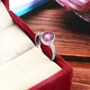 Könnte beinhalten: Ein silberner Ring mit einem großen, runden, purpurroten Edelstein. Der Edelstein hat ein sternförmiges Muster. Der Ring wird in einer roten Samt-Schmuckschatulle präsentiert.
