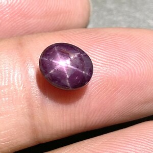 Könnte beinhalten: Ein ovaler Sternrubin-Edelstein im Cabochon-Schliff mit einem tiefen violett-roten Farbton. Der Stein weist einen ausgeprägten sechsstrahligen Sterneffekt auf, bekannt als Asterismus, der als weißer Lichtreflex die Oberfläche durchzieht.