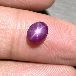 Könnte beinhalten: Ein tief magentafarbener Sternrubin-Edelstein mit einem glatten, polierten Cabochon-Schliff. Der oval geformte Stein zeigt einen ausgeprägten sechsstrahligen Sterneffekt, bekannt als Asterismus, der als heller weißer Lichtreflex erscheint.