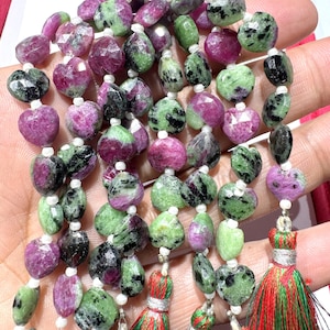 Könnte beinhalten: Halskette aus Edelsteinperlen in Grün, Rosa und Schwarz. Die Perlen sind auf weißem Faden aufgefädelt und mit Quasten in Rot, Grün und Gold versehen. Die Perlen haben eine Herzform. Die Halskette ist ca. 40 cm lang.