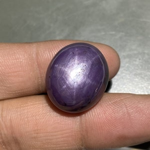 May include: A polished, oval-shaped, purple gemstone with a six-ray star pattern. The gem is a deep violet color with a smooth, reflective surface. The star effect is visible across the surface of the stone.