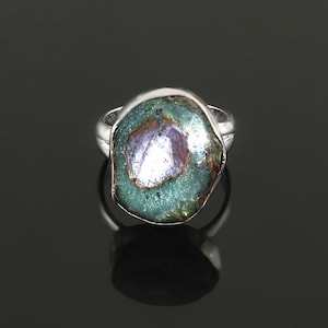 Könnte beinhalten: Ein Silberring mit einem großen, unregelmäßig geformten Stein. Der Stein hat eine teal-grüne Außenschicht mit einem zentralen, facettenreichen, lavendelfarbenen Kristall. Der Ring hat ein glattes, poliertes Band, das einen Kontrast zum strukturierten Stein bildet.