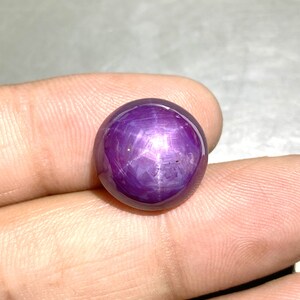 Peut inclure: Une pierre précieuse rubis étoilé de forme ronde et taillée en cabochon, présentant une teinte rouge-violet profond. La pierre affiche un effet d'étoile à six branches distinct, appelé astérisme, sous forme de motif lumineux croisé.