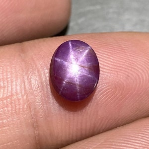 Könnte beinhalten: Ein ovaler Sternrubin-Edelstein mit einem tiefen purpurroten Farbton und einem ausgeprägten sechsstrahligen Sternmuster, dem sogenannten Asterismus, das das Licht von der Mitte der glatten, polierten Oberfläche des Cabochons reflektiert.