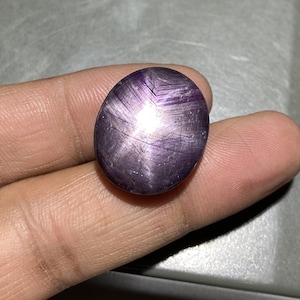 May include: A polished, oval-shaped, purple gemstone with a star-like pattern radiating from the center. The stone has a smooth surface with visible internal lines and a bright, reflective star effect. The gem is held in a hand.