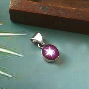Puede incluir: Colgante de plata con una piedra preciosa de rubí estrella. El rubí ovalado es de color rojo púrpura intenso y presenta un efecto de estrella de seis rayos. El colgante tiene un diseño sencillo y elegante.