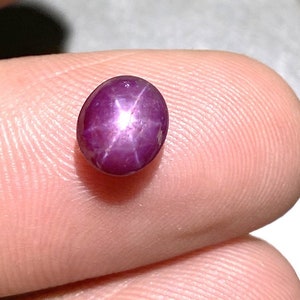 Könnte beinhalten: Ein polierter, oval geformter Cabochon-Edelstein mit einem tiefen Magenta-Violett-Ton. Der Stein weist einen ausgeprägten sechsstrahligen Sterneffekt auf, bekannt als Asterismus, der als heller weißer Lichtreflex auf der glatten Oberfläche erscheint.