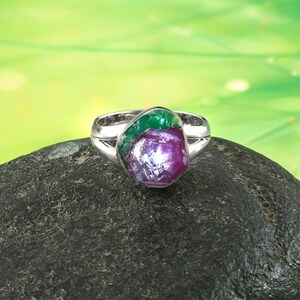 Pode incluir: Um anel de prata com uma grande pedra preciosa de forma irregular. A pedra apresenta tons de vermelho profundo, roxo e verde, contrastando com a banda de prata. O anel est&aacute; sobre uma superf&iacute;cie escura e texturizada.