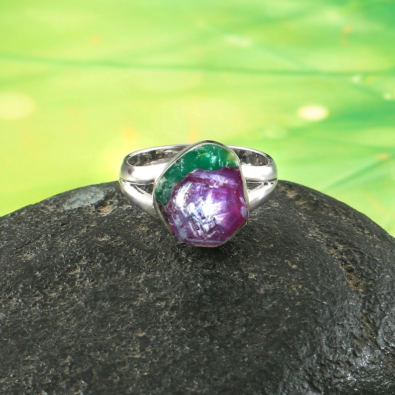 Pode incluir: Um anel de prata com uma grande pedra preciosa de forma irregular. A pedra apresenta tons de vermelho profundo, roxo e verde, contrastando com a banda de prata. O anel est&aacute; sobre uma superf&iacute;cie escura e texturizada.