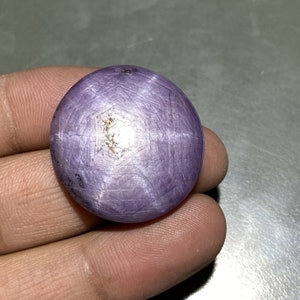 May include: A round, polished, lavender-colored gemstone with a star-like pattern across its surface. The stone has a smooth, reflective finish and a small, light-colored inclusion near the center. The gemstone is held in a hand.
