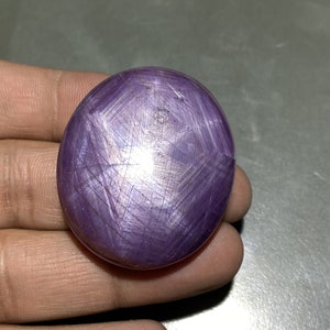May include: Oval-shaped, polished purple gemstone with a star-like pattern. The gemstone has a smooth, reflective surface with intersecting lines creating a star effect. The stone is held in a hand, showcasing its size and color.