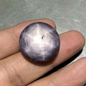 May include: A round, polished, star sapphire gemstone. The gem is a deep purple color with a six-rayed star pattern visible on its surface. The stone has a smooth, reflective finish.