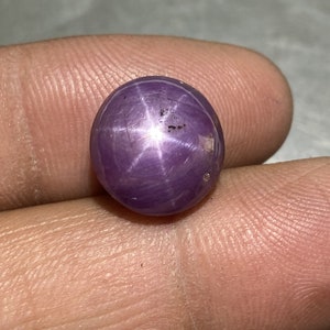 Könnte beinhalten: Ein runder, im Cabochon-Schliff gefertigter Sternrubin-Edelstein mit einem tiefen violett-rosa Farbton. Der Stein zeigt einen ausgeprägten sechsstrahligen Asterismus-Effekt, der als helles, weißes Sternmuster auf der polierten Oberfläche erscheint.