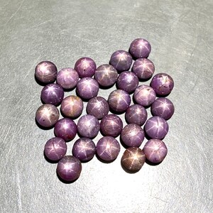 Op de afbeelding: Een verzameling ronde, gepolijste edelstenen in stervorm, in verschillende tinten paars. Elke edelsteen toont een zesstralig sterpatroon. De edelstenen liggen op een grijze ondergrond, waardoor hun unieke structuren worden benadrukt.