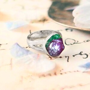Pode incluir: Um anel de prata com uma gema de rubi bruta, exibindo tons verdes e roxos. A banda do anel tem um design dividido, e a gema &eacute; o ponto focal. O anel est&aacute; sobre um fundo macio e padronizado.