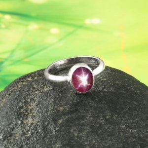Könnte beinhalten: Ein silberner Ring mit einem ovalen Sternrubin-Edelstein. Der Rubin hat eine tiefrote Farbe mit einem sechsstrahligen Sternmuster. Der Ring liegt auf einem dunkelgrauen Stein vor einem verschwommenen grün-gelben Hintergrund.