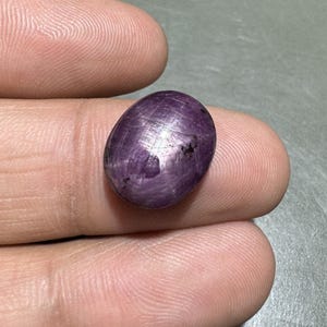 Könnte beinhalten: Ein polierter, oval geformter, purpurroter Edelstein. Der Edelstein hat eine glatte Oberfläche mit sichtbaren inneren Linien und einem dunklen Fleck. Der Stein wird zwischen zwei Fingern gehalten, was seine Farbe und Form zeigt.