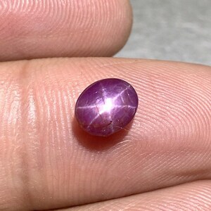 Könnte beinhalten: Ein im Cabochon-Schliff gefertigter Sternrubin-Edelstein mit einem tiefen Magenta-Violett-Ton. Der Stein weist einen ausgeprägten sechsstrahligen Asterismus-Effekt auf, bei dem ein helles weißes Sternmuster aus der Mitte der polierten Oberfläche strahlt.