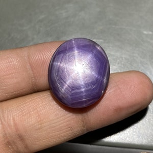 May include: A polished, oval-shaped, purple gemstone with a star-like effect. The gem has a smooth, reflective surface with intersecting lines creating a six-pointed star. The stone is held in a hand, showcasing its size and color.