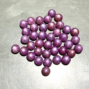 Op de afbeelding: Een verzameling ronde, gepolijste edelstenen in verschillende tinten paars en roze. Elke edelsteen vertoont een zesstralig sterpatroon. De edelstenen zijn gerangschikt op een grijze ondergrond, wat een aantrekkelijke opstelling creëert.