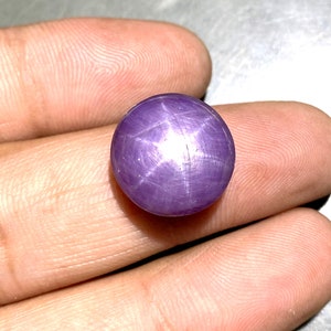 Könnte beinhalten: Ein runder, im Cabochon-Schliff gehaltener violetter Sternsaphir-Edelstein mit einem ausgeprägten sechsstrahligen Asterismus-Effekt, der von der Mitte des Steins ausgeht. Die Oberfläche des Edelsteins ist glatt und poliert und zeigt einen tiefvioletten Farbton.