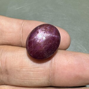 Könnte beinhalten: Ein polierter, oval geformter Edelstein mit tief burgunderroter Farbe und einer schimmernden, irisierenden Oberfläche. Der Stein hat eine glatte, abgerundete Form, und das Licht fängt seine Oberfläche ein und erzeugt ein Farbspiel. Der Edelstein wird in einer Hand gehalten.