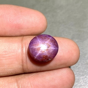 Puede incluir: Una gema de rubí estrella redonda y pulida. La gema es de color rojo púrpura intenso con un patrón de estrella de seis rayos visible en su superficie. La piedra se sostiene entre dos dedos.