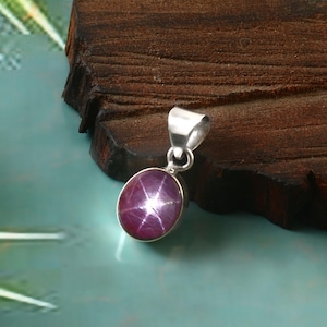 Könnte beinhalten: Silberanhänger mit einem großen, ovalen Sternrubin-Edelstein. Der Rubin hat eine tiefrot-violette Farbe mit einem sechsstrahligen Sternmuster. Der Anhänger hat ein schlichtes, elegantes Design, perfekt für den Alltag.