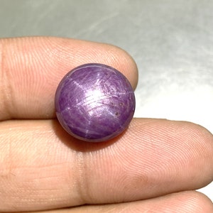 May include: A round, cabochon-cut star ruby gemstone with a deep purplish-red hue. The surface displays a distinct six-rayed star effect, known as asterism, created by light reflecting off internal needle-like inclusions within the polished stone.