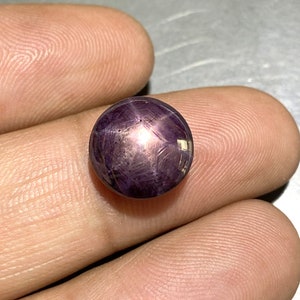 May include: A round, polished, purple gemstone with a star-like pattern. The gem is held between fingers, showcasing its color and the star effect created by light reflecting off the internal structures. The gem is a cabochon cut, with a smooth, rounded surface.