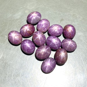 Könnte beinhalten: Eine Ansammlung von zwölf ovalen, im Cabochon-Schliff bearbeiteten Sternrubinen in verschiedenen Schattierungen von tiefem Magenta und Violett. Jeder Edelstein zeigt einen deutlichen sechsstrahligen Sterneffekt, Asterismus genannt.