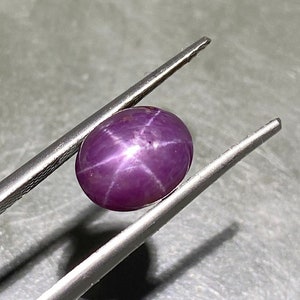 Könnte beinhalten: Ein ovaler, tief magentafarbener Sternrubin-Edelstein mit einem ausgeprägten sechsstrahligen Asterismus-Muster auf seiner glatten, polierten Cabochon-Oberfläche. Der Edelstein wird sicher zwischen den beiden Metallspitzen einer Pinzette gehalten.