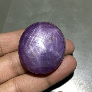 May include: A round, polished, purple gemstone with a star-like pattern across its surface. The gemstone is held in a hand, showcasing its color and the way light reflects off its facets. The stone's surface has a textured appearance.