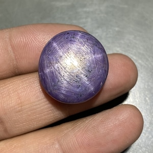 May include: A round, polished, purple gemstone with a star-like pattern. The gem has a smooth surface and a subtle sheen, with a central area of lighter color. The star effect is created by intersecting lines across the surface.