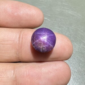 Könnte beinhalten: Ein runder, polierter, violetter Edelstein mit einem sternartigen Muster auf der Oberfläche. Der Edelstein wird zwischen zwei Fingern gehalten, wodurch seine Farbe und Textur hervorgehoben werden. Der Edelstein scheint ein Sternsaphir oder ein ähnlicher Edelstein zu sein.