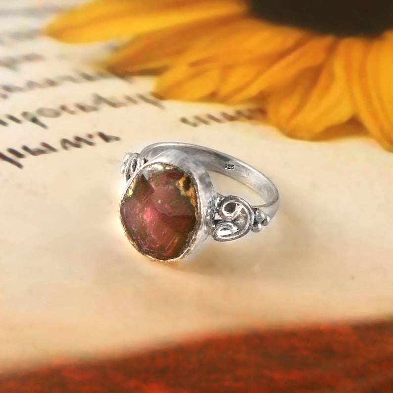 May include: A silver ring featuring an oval, reddish-brown gemstone. The ring has decorative swirls on the sides. It is set against a background of a sunflower and handwritten text.