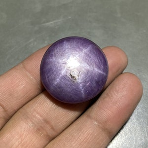 May include: A round, polished, purple gemstone with a six-pointed star pattern. The gem has a smooth surface and a slight imperfection near the center of the star. The stone is held in a hand.