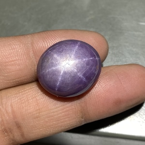 May include: A polished, oval-shaped, purple star sapphire gemstone. The gem displays a six-rayed star pattern across its surface. The stone is held between two fingers, showcasing its color and luster. The gem is a deep violet hue.