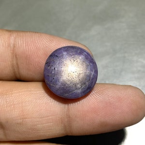May include: A round, cabochon-cut purple star sapphire gemstone featuring a distinct six-rayed star effect centered on its surface. The stone displays a translucent to opaque quality with visible dark inclusions and a smooth, polished dome finish.
