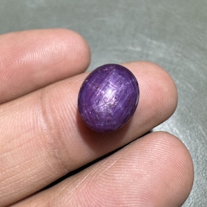 Könnte beinhalten: Ein polierter, ovaler, violetter Edelstein mit Sterneneffekt. Der Edelstein wird zwischen den Fingern gehalten, wodurch seine Farbe und Textur sichtbar werden. Der Stein scheint ein Sternrubin, eine Art Korund, zu sein.