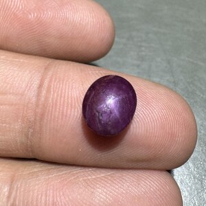 Könnte beinhalten: Ein polierter, oval geformter, tiefvioletter Edelstein. Der Edelstein hat einen sternartigen Effekt auf seiner Oberfläche. Der Stein wird zwischen den Fingern gehalten, wodurch seine Farbe und Form zur Geltung kommen. Dieser Edelstein könnte in Schmuck verwendet werden.