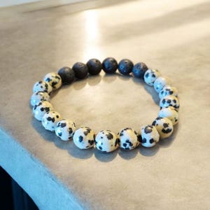 Peut inclure: Bracelet de perles composé de perles de lave noire et de jaspe dalmatien. Les perles de jaspe dalmatien sont blanches avec des taches noires. Le bracelet est monté sur un cordon élastique.